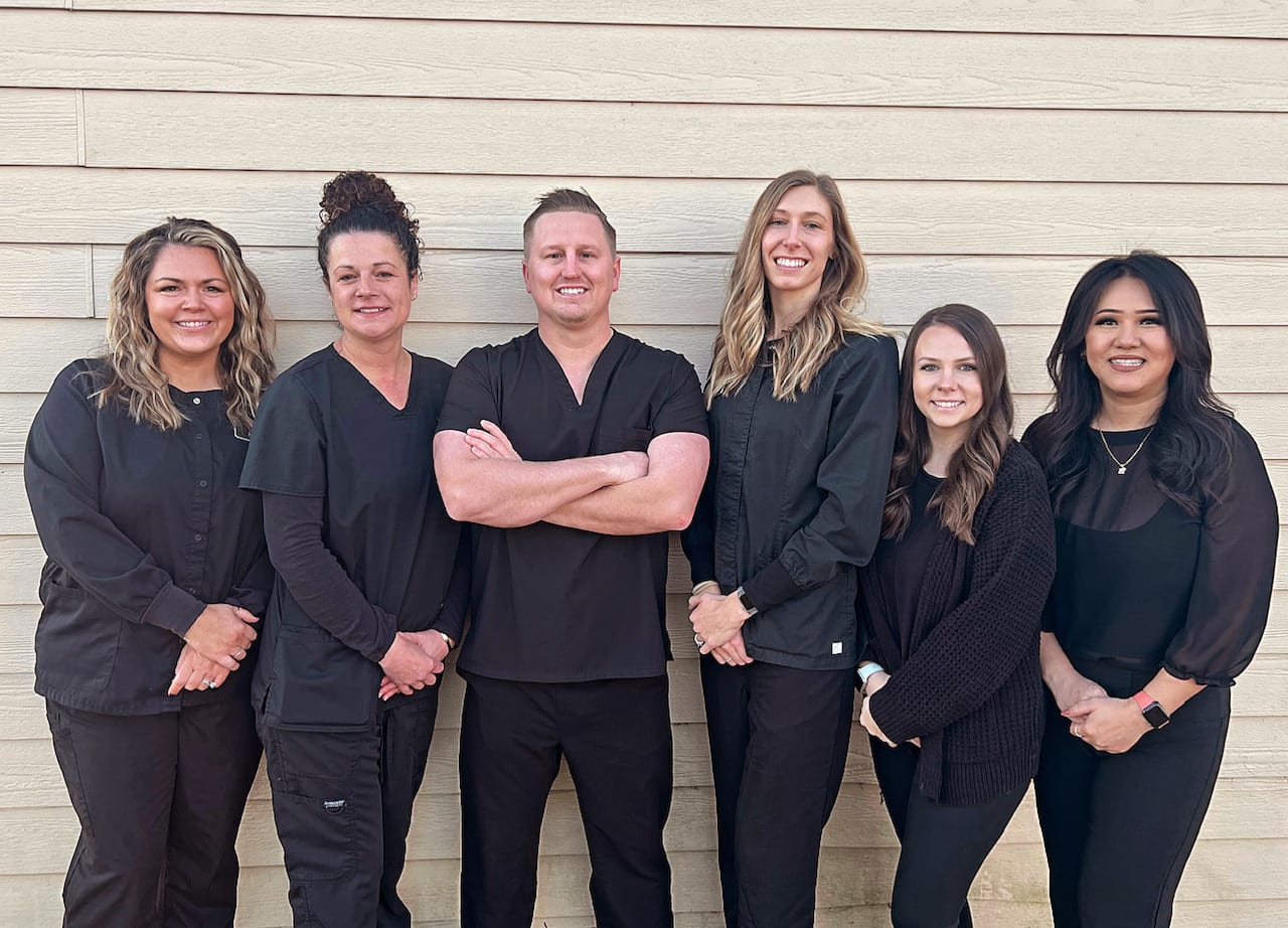 Six dental team members in black scrubs smiling outside office building.